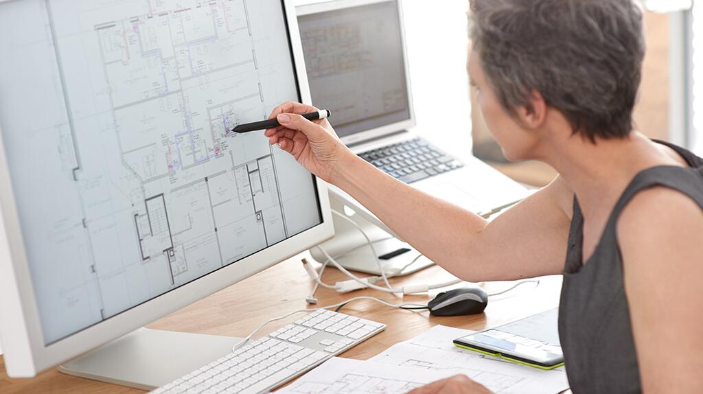 Vrouw wijst met pen naar bouwtekening op computerscherm.