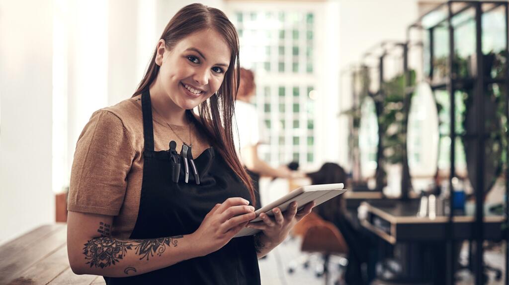 Vrouw met een schort en tablet, glimlachend in een moderne salon.