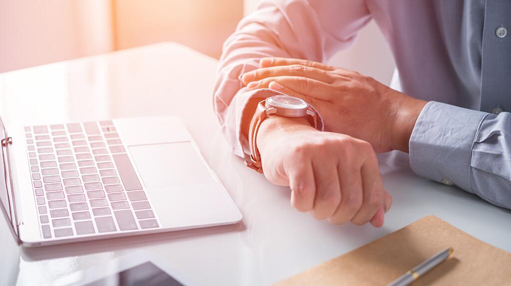 Man in hemd kijkt naar horloge, laptop op tafel.
