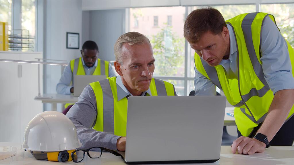 Twee mannen in veiligheidshesjes werken aan laptop in kantooromgeving.