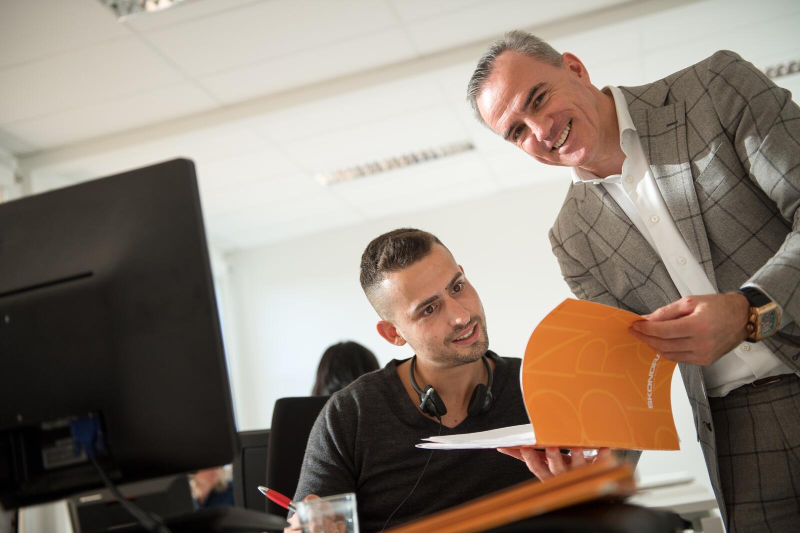 Man met map geeft uitleg aan iemand bij een computerscherm in kantooromgeving.