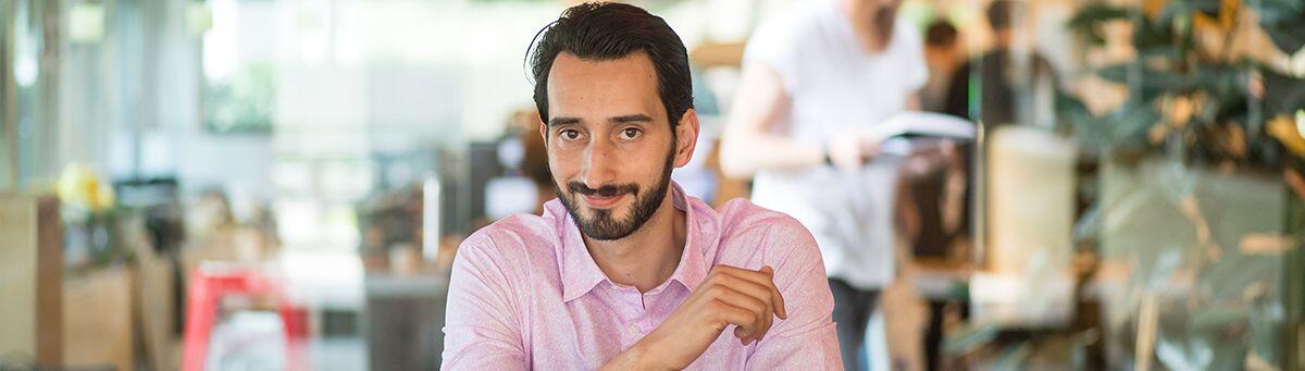 Man in roze overhemd glimlacht in een drukke kantoorsetting.