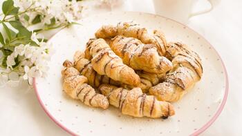 Croissants op een wit bord, gedecoreerd met bloemen en een kopje op de achtergrond.
