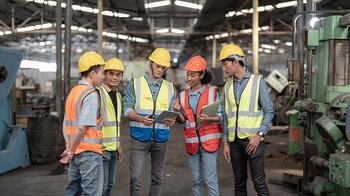 Groep arbeiders met helmen en hesjes bespreekt iets op tablet in fabriekshal.