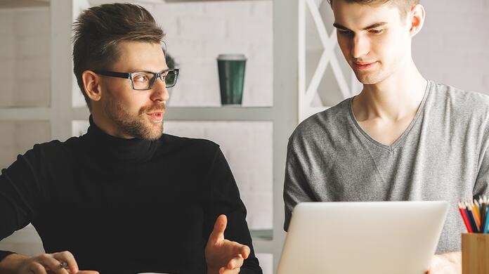 Twee mannen praten en werken op een laptop in een kantooromgeving.