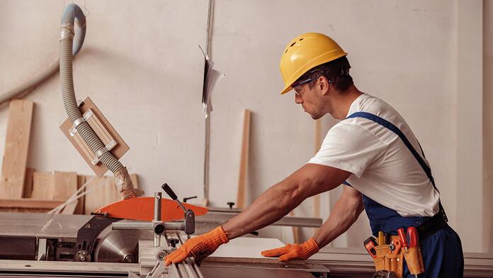 Man met gele helm zaagt hout in werkplaats.