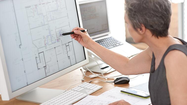 Vrouw wijst met pen naar bouwtekening op computerscherm.