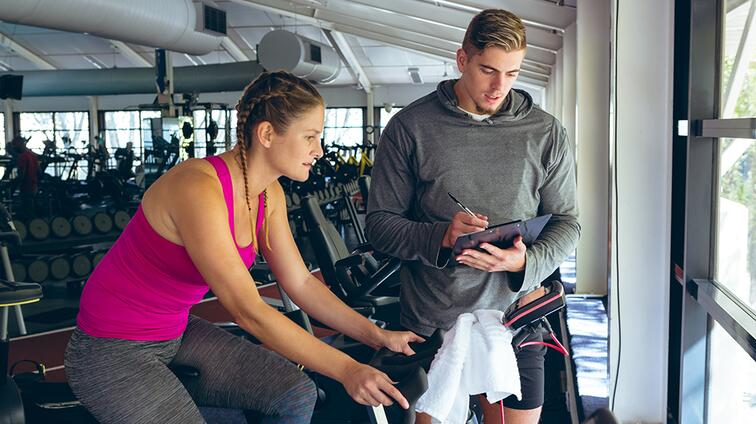 Vrouw op hometrainer luistert naar man met clipbord in sportschool.