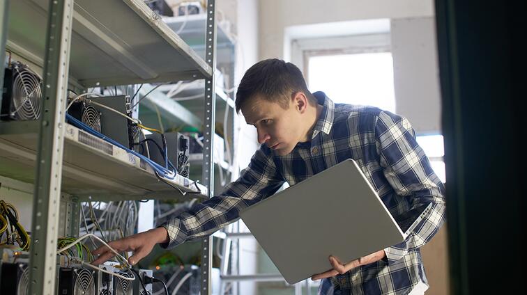Persoon in serverruimte met laptop, kijkend naar apparatuur.