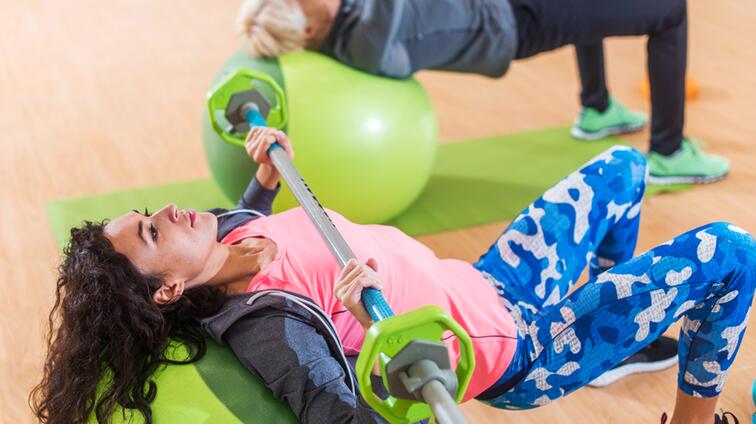 Vrouw tilt een halter boven haar hoofd, liggend op een groene mat in een fitnessruimte.